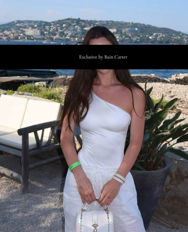 Aurora posing in a white one-shoulder dress with a coastal background.