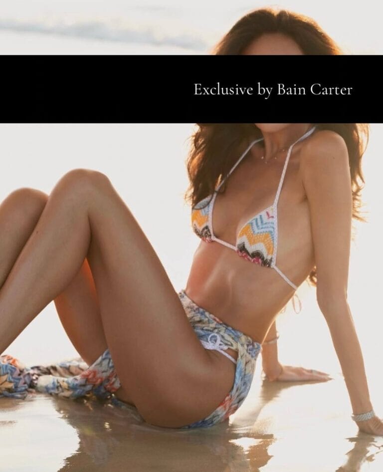 Karina wearing a patterned colorful bikini reclining on the wet sand at the beach.