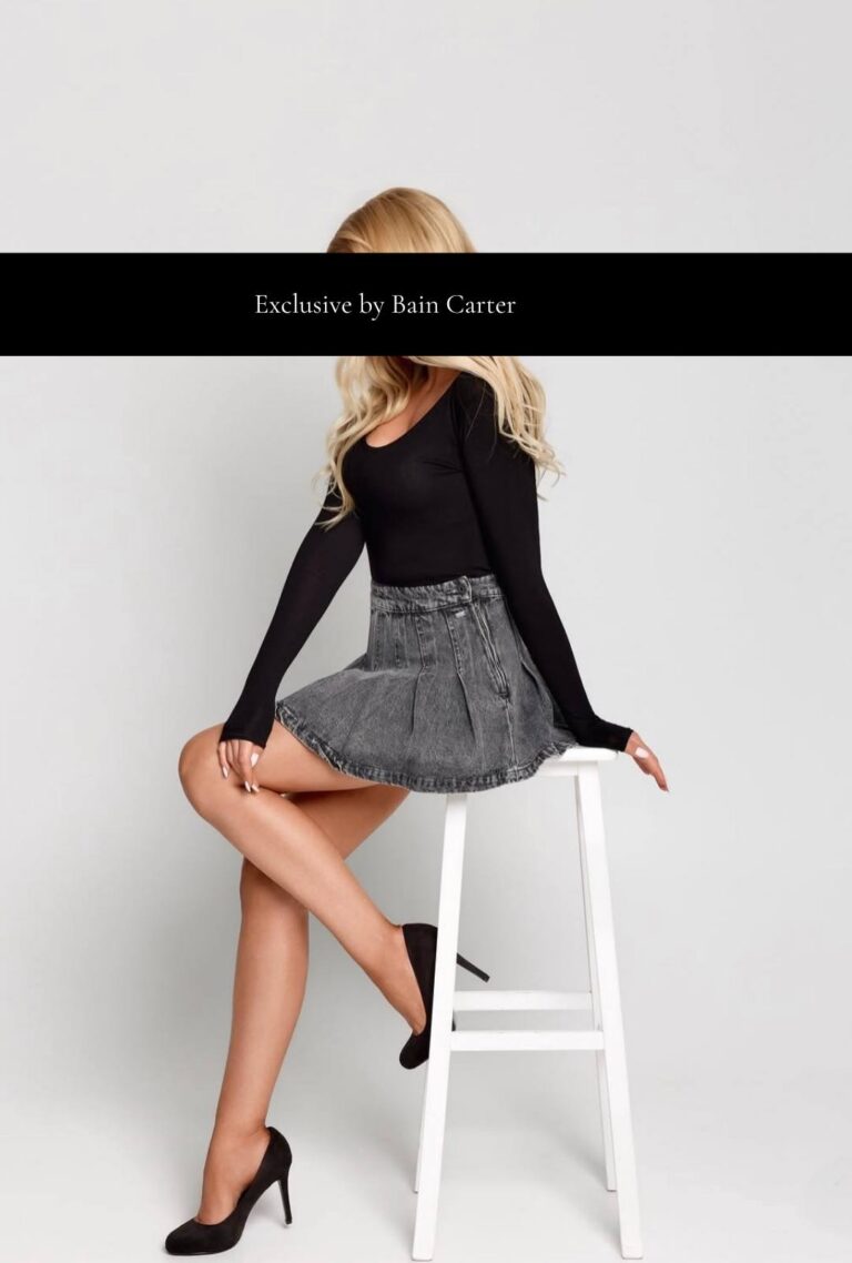 Amelie sitting on a stool wearing a black top, grey skirt and heels