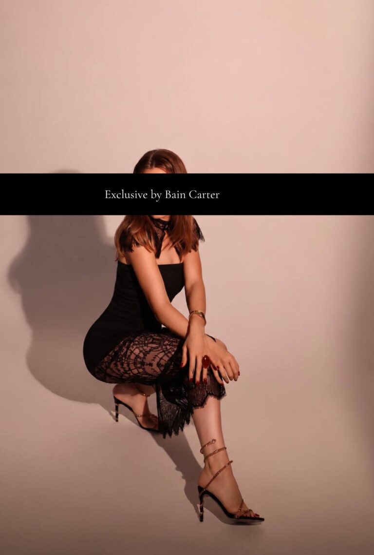 Violetta seated in a black dress with lace panels and strappy heels